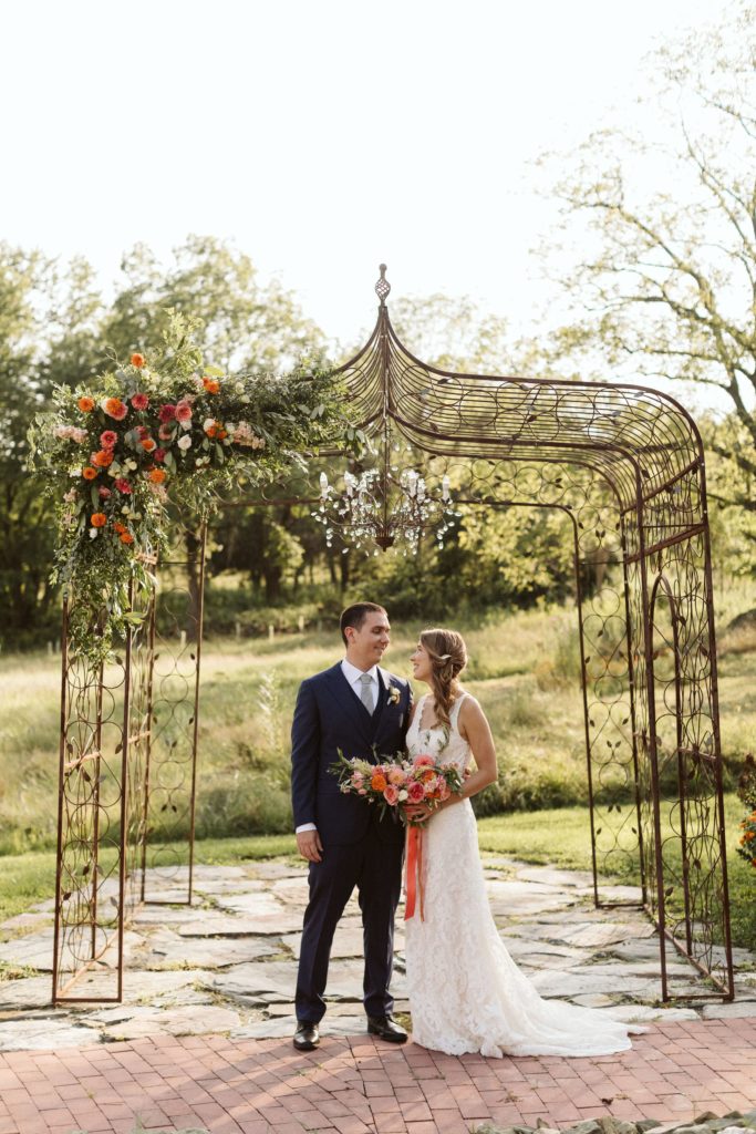 A couple stands under an arch decorated with coral, pink and orange flowers at Zigbone Farm Retreat