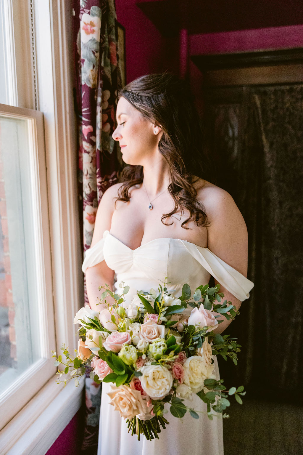 Bride stands by the window while holding a peach and pink bridal bouquet with roses and eucalyptus at Iron Gate Restaurant, Washington DC